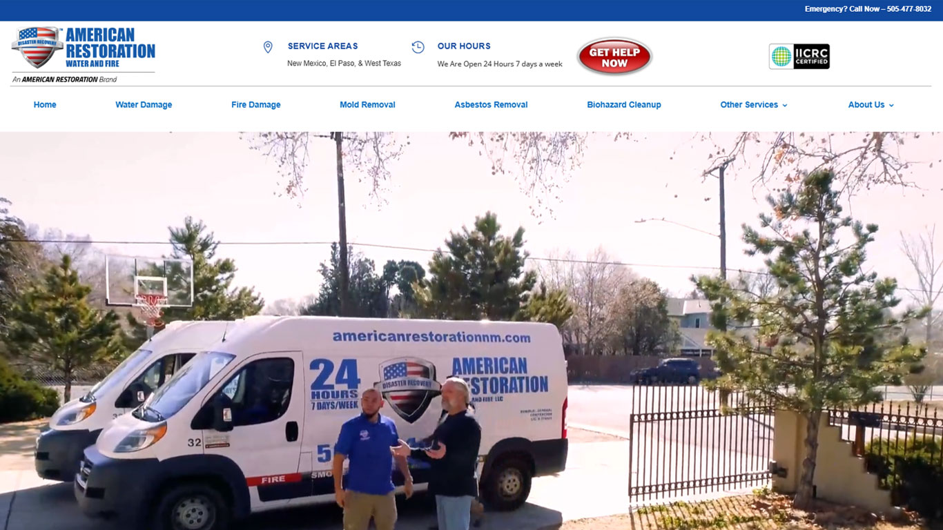 White restoration van with American Restoration branding parked in a suburban driveway, two men standing beside it among trees and a gated yard.