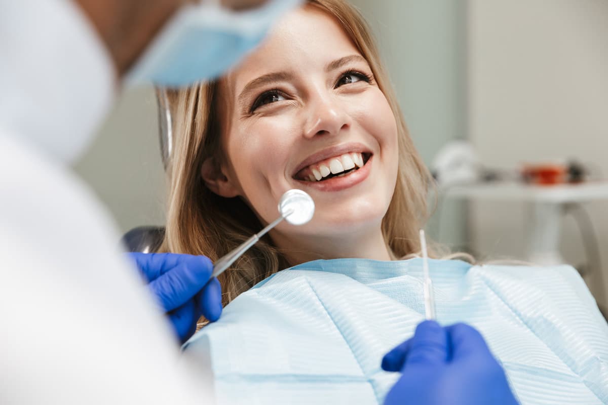 Woman in a dental chair smiling as a dentist in blue gloves uses a mirror to examine her teeth.